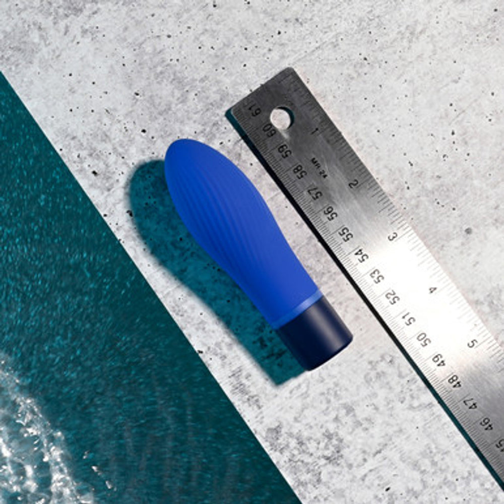A blue silicone bullet vibrator lies next to a metal ruler on a concrete surface.