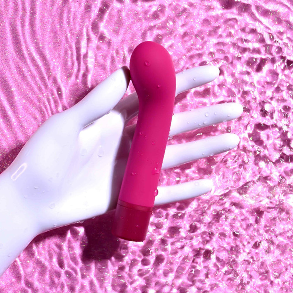 A pink G-spot vibrator rests on a white mannequin hand in shimmering pink water.