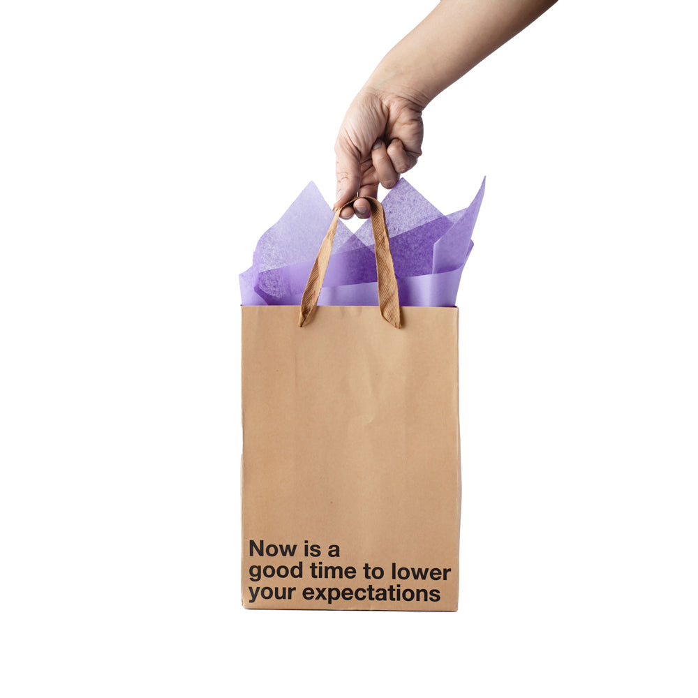 A kraft paper gift bag with purple tissue paper and the text "Now is a good time to lower your expectations".
