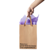 A kraft paper gift bag with purple tissue paper and the text "Now is a good time to lower your expectations".