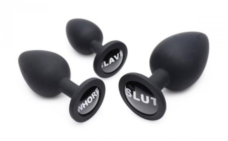 Three black silicone anal plugs with "Slave", "Whore", and "Slut" inscribed on them.