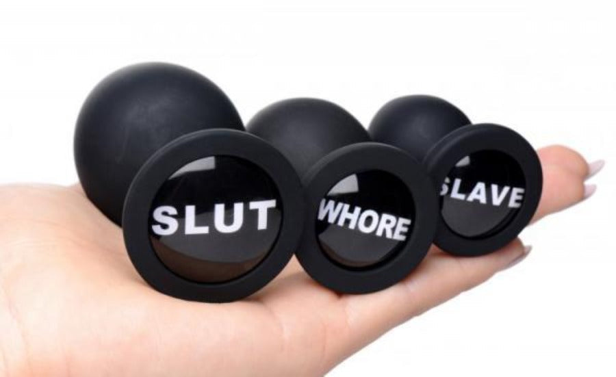 Black silicone anal plugs with "SLUT", "WHORE", and "SLAVE" inscribed on them.