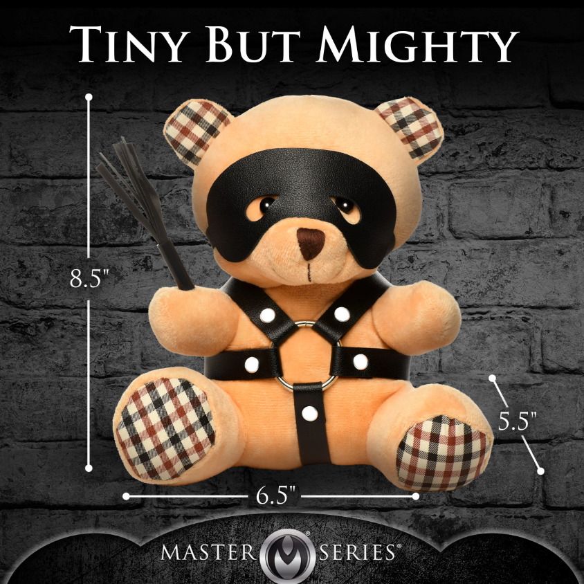 A tan teddy bear wearing a black eye mask and faux leather harness, holding a mini flogger.
