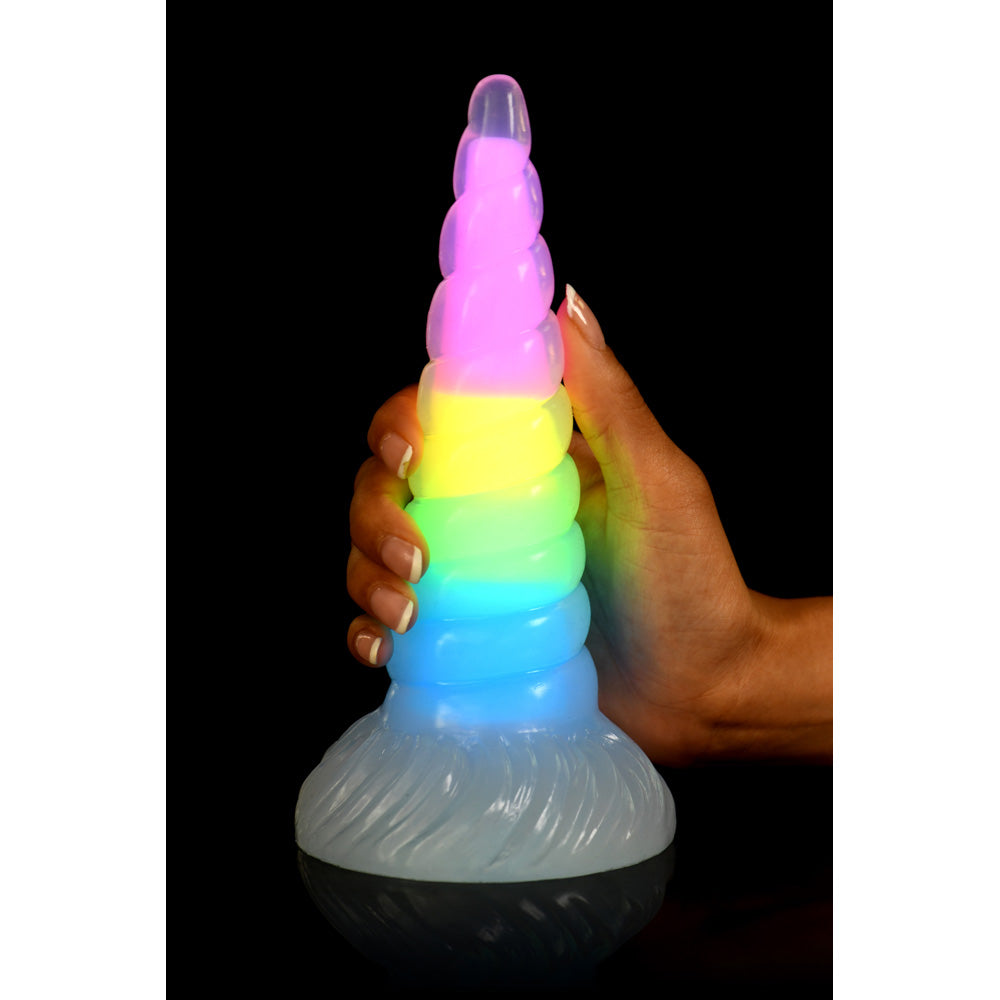 A hand holds a rainbow-colored, glowing unicorn horn dildo.