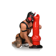 A muscular man in a black leather harness and red dog hood kneels holding a giant red canine fantasy dildo with suction cup base.