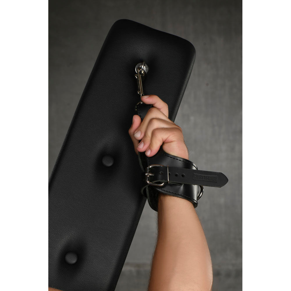 A hand in a black leather cuff is attached to a metal ring on a black padded surface.