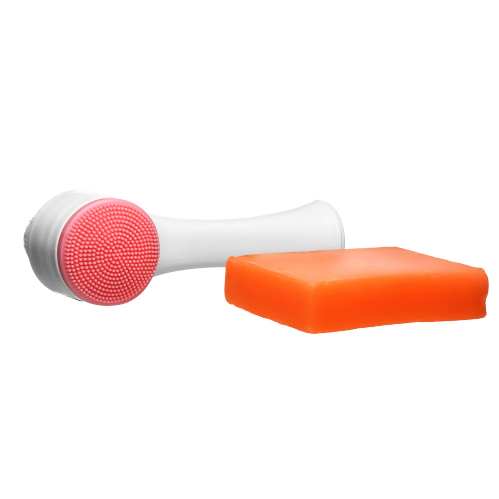 A white exfoliating facial brush with a pink silicone head and a bar of orange soap.