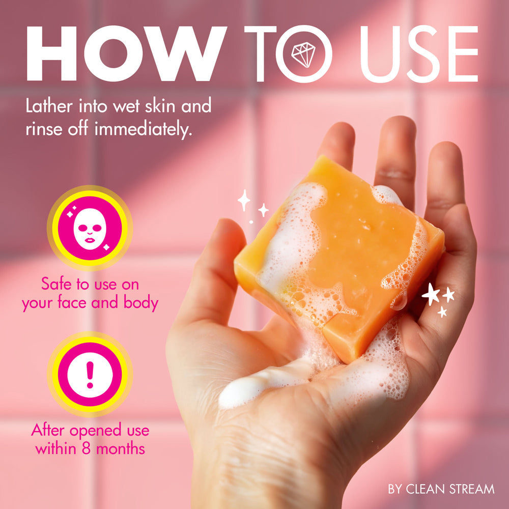 A hand holds a bar of orange soap with white lather against a pink tiled background.