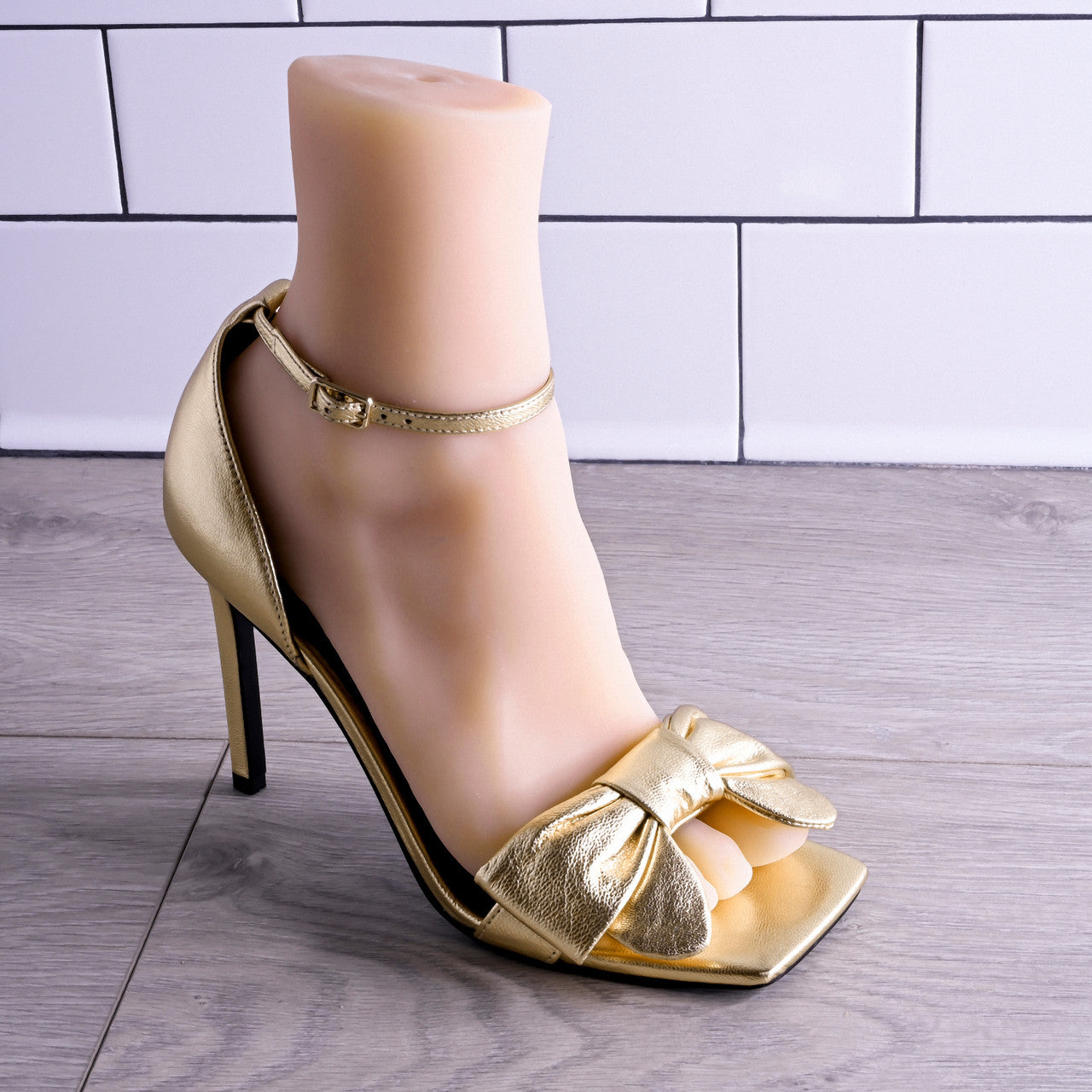 Gold stiletto sandal with bow detail on a foot mannequin.