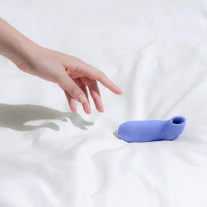 A purple silicone suction stimulator on a white sheet with a hand reaching towards it.
