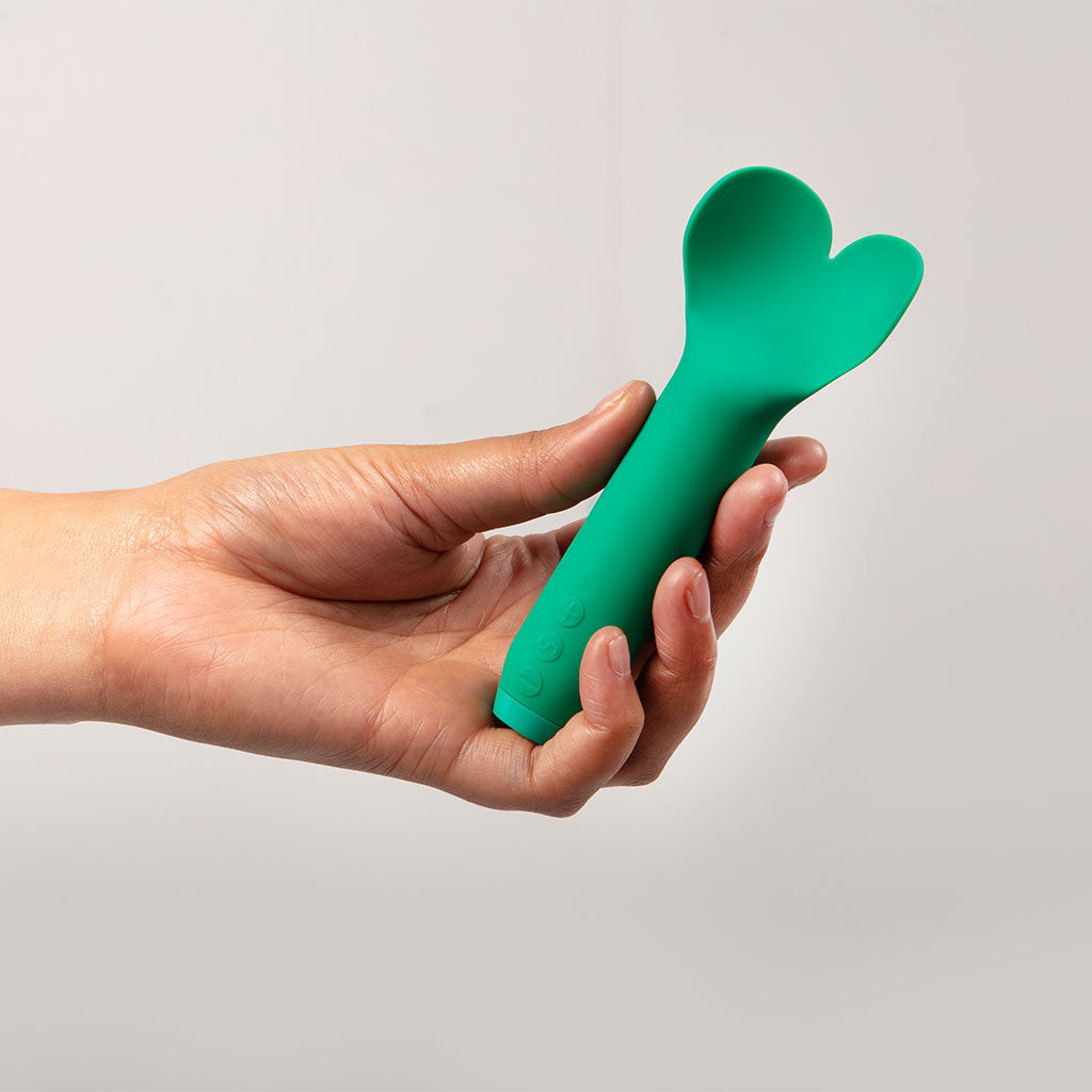 A hand holds a green silicone vibrator with a heart-shaped tip.