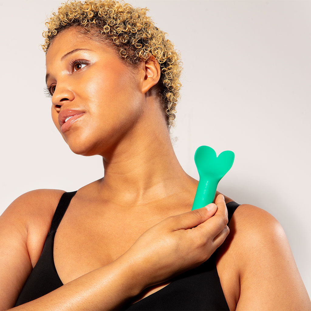 A woman holds a green heart-shaped vibrator to her shoulder.