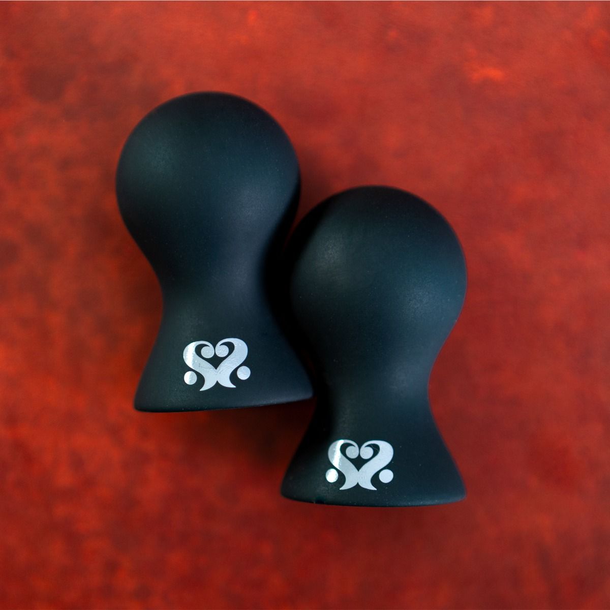 Two black nipple suckers with a silver heart-shaped logo on a red background.