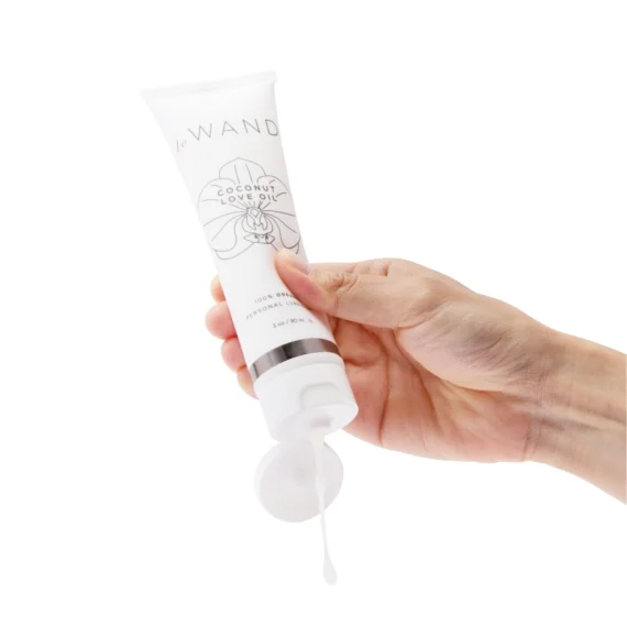 A hand squeezes a tube of Le Wand Coconut Love Oil, with a drop of white lubricant falling from the open cap.