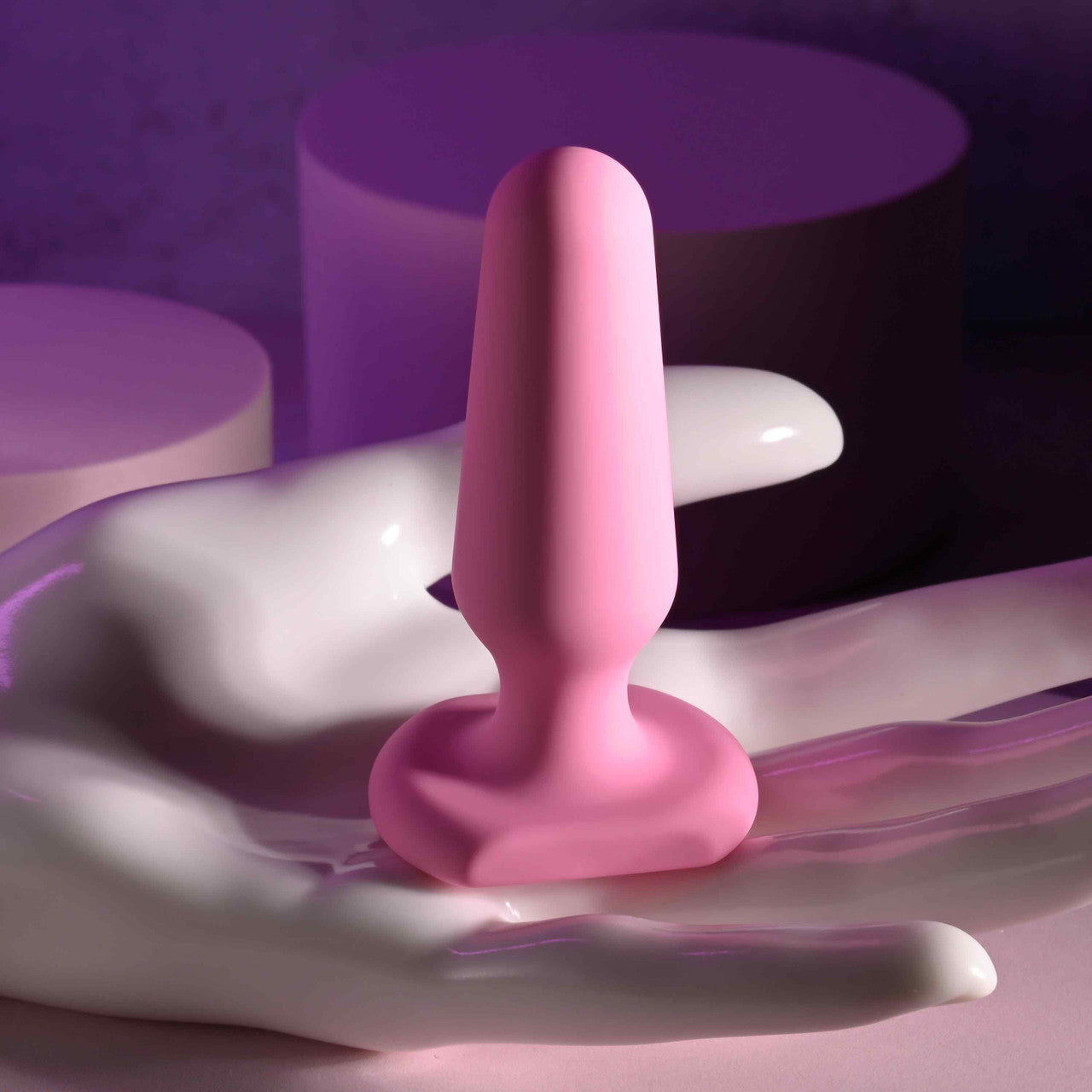 A pink silicone anal plug with a flared base rests on a white hand-shaped sculpture.