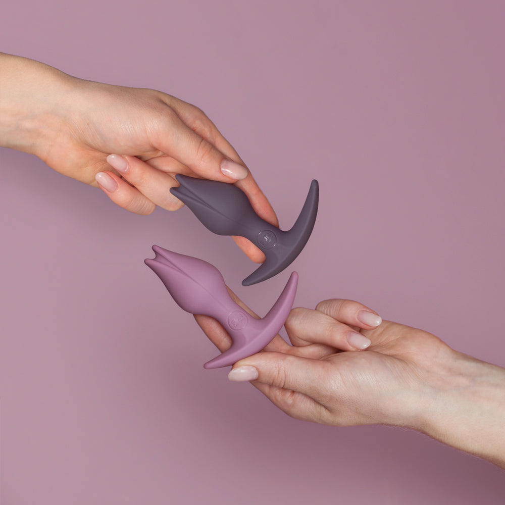 Two hands hold purple and grey anal plugs with asymmetrical bases.