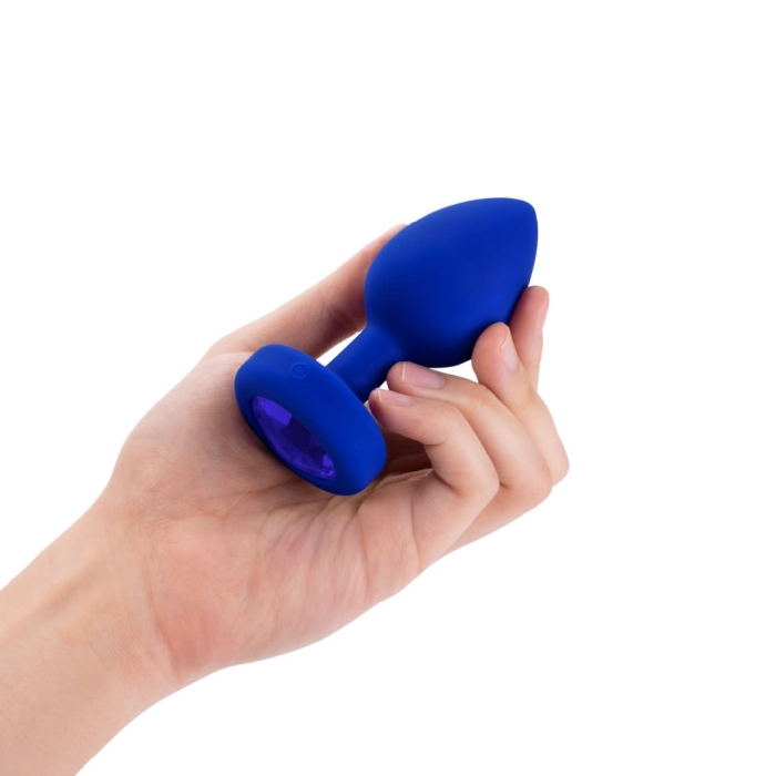 A hand holds a navy blue silicone vibrating anal plug with a purple jewel accent.