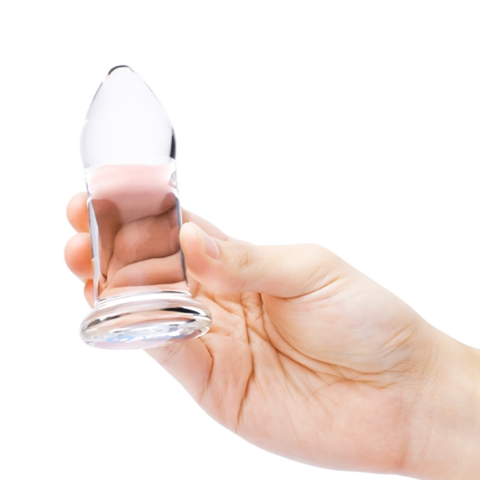 A hand holds a clear glass anal dilator with a tapered tip and flared base.