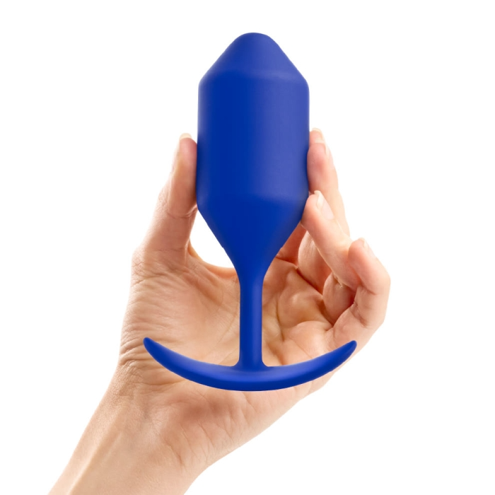 A hand holds a navy blue weighted silicone anal plug with a flared base.