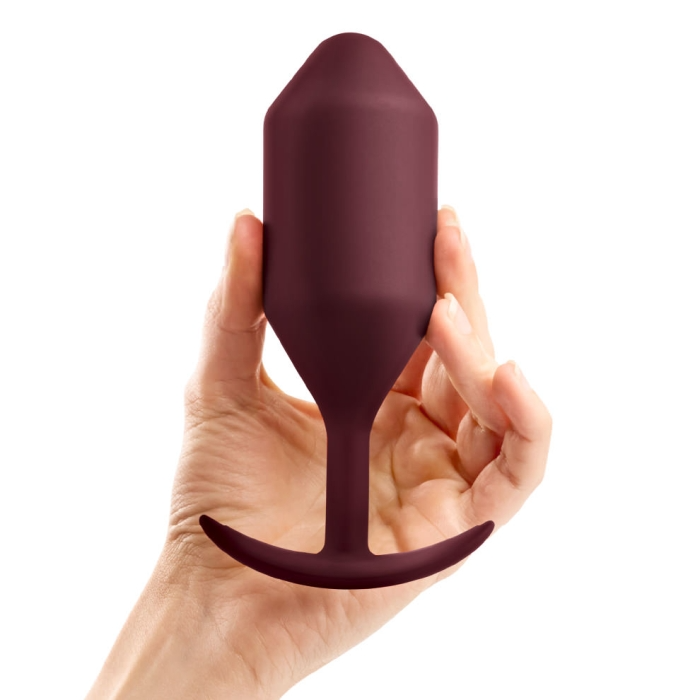 A hand holds a dark red, weighted silicone anal plug with a flared base.