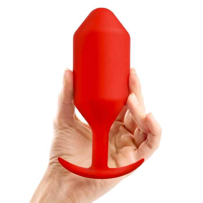 A hand holds a red weighted silicone anal plug with a tapered tip and T-shaped base.