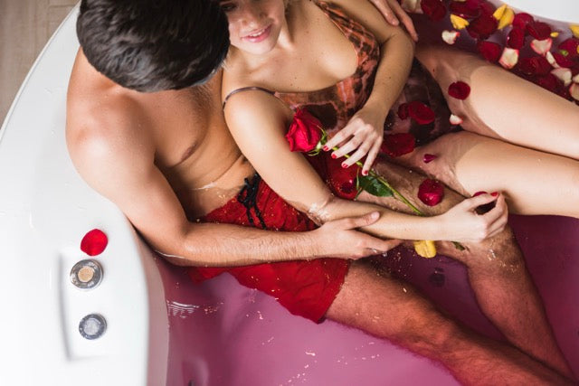 Two people in a bathtub with rose petals and red roses, surrounded by rose petals.