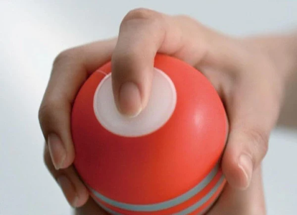 A hand holds a red Tenga vacuum cup masturbator with a white button.