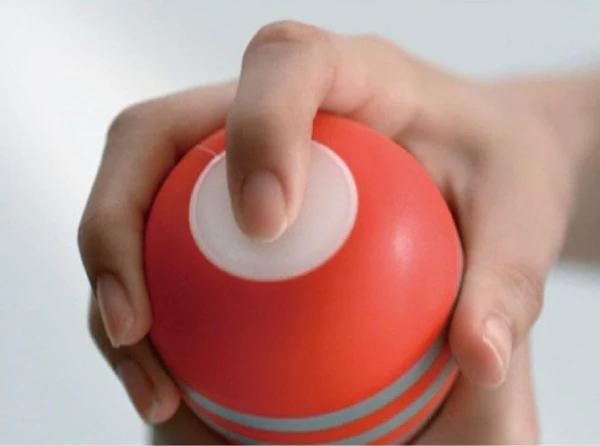 A hand grips a red Tenga masturbator with a white button.