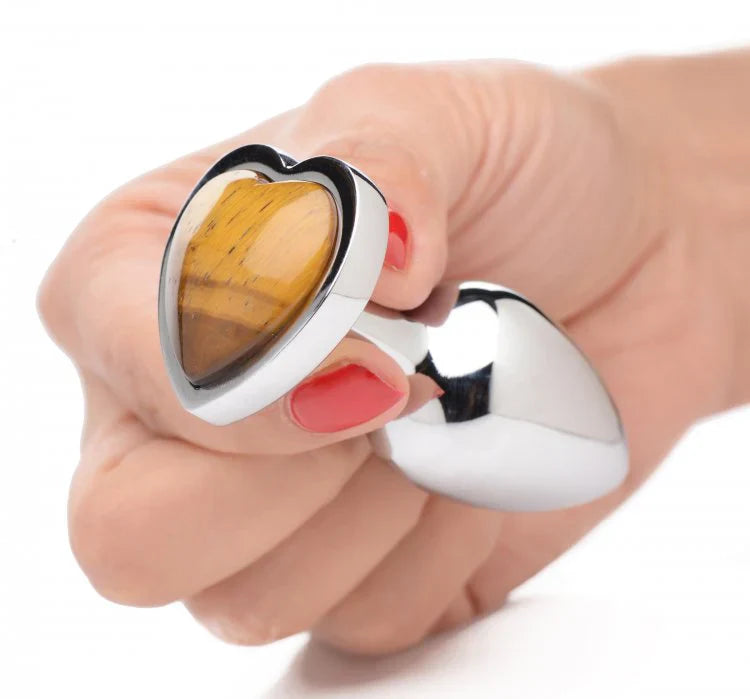 A hand holds a heart-shaped anal plug with a tiger's eye gemstone and aluminum alloy base.