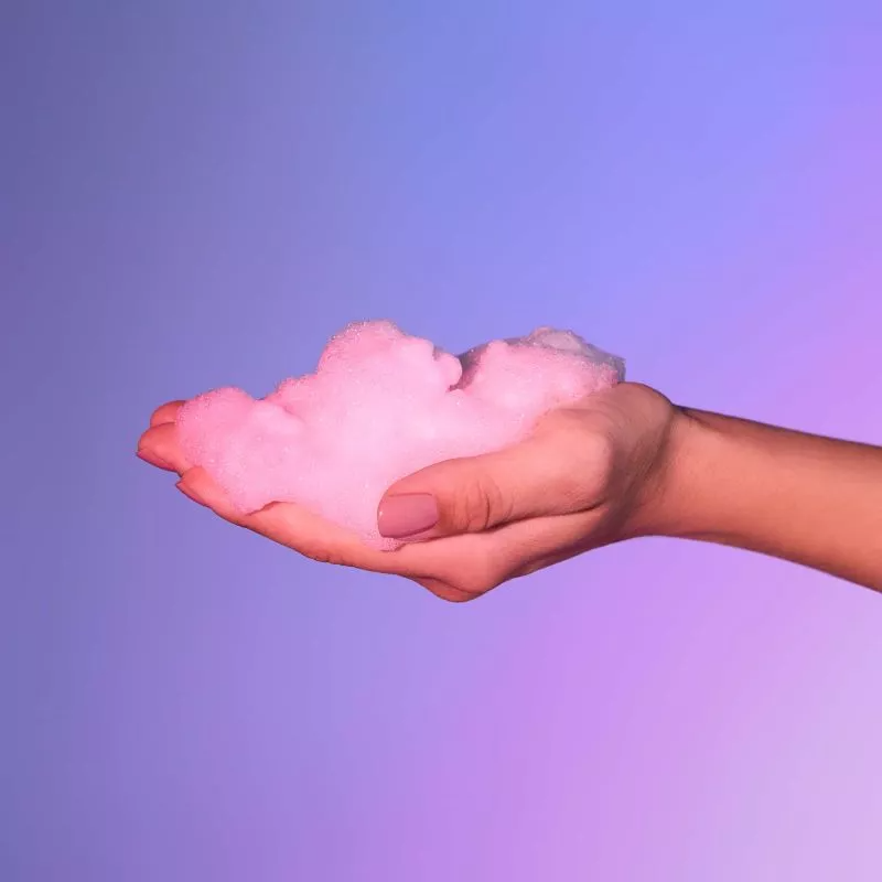 A hand holds pink cherry blossom scented massage foam against a purple and blue gradient background.