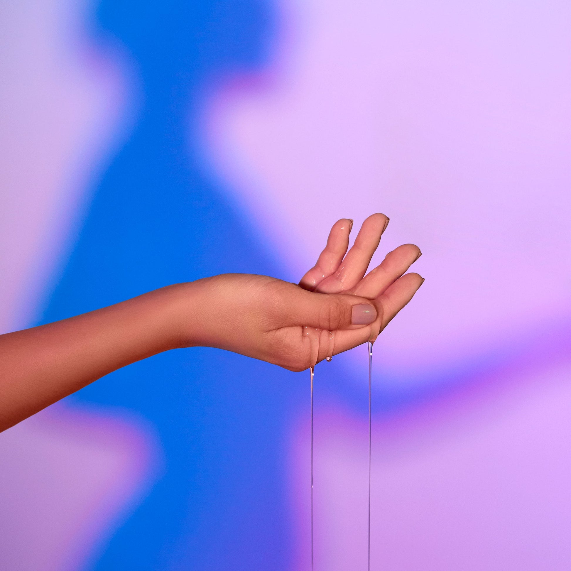 Hand with warming massage oil dripping from fingers against a purple and blue background.