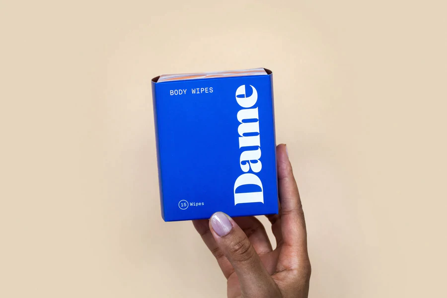 A hand holds a blue box of Dame body wipes with white text.