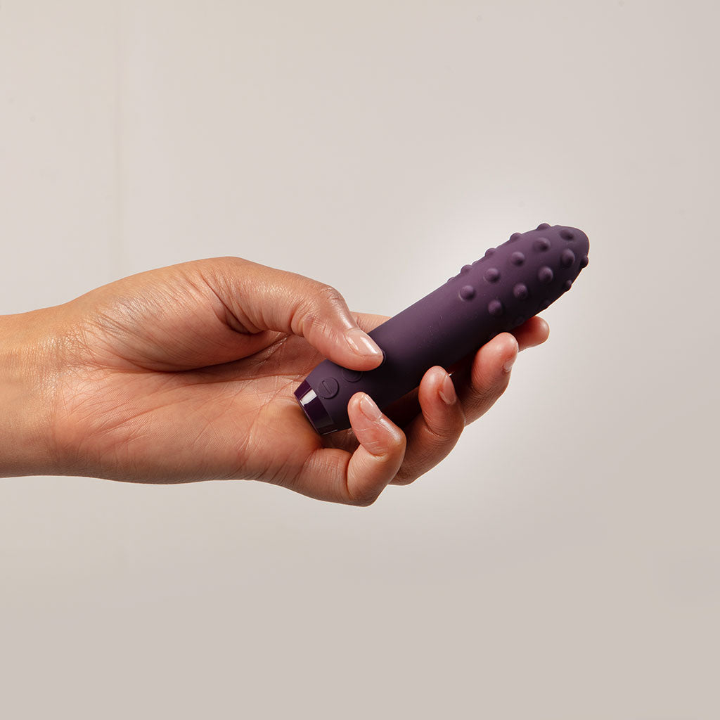A hand holds a purple silicone bullet vibrator with raised bumps.