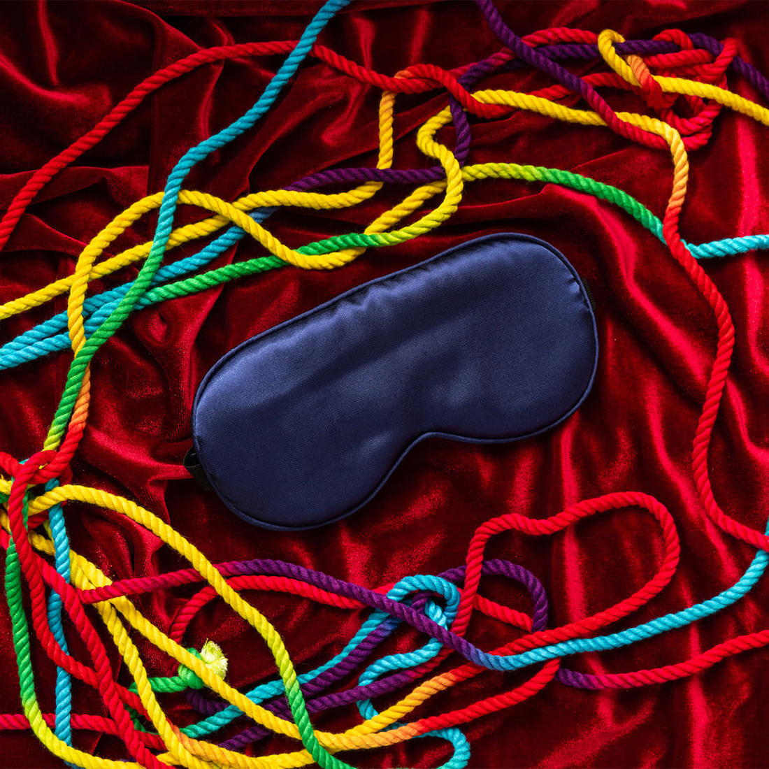 Midnight blue satin sleep mask and bondage blindfold on red velvet with colorful ropes.