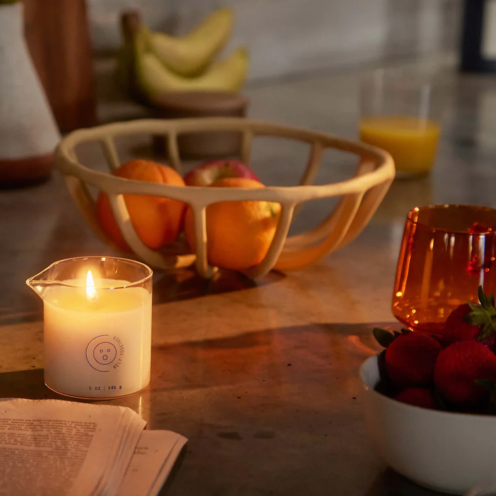 A lit massage oil candle with a pouring spout sits on a counter next to fruit.
