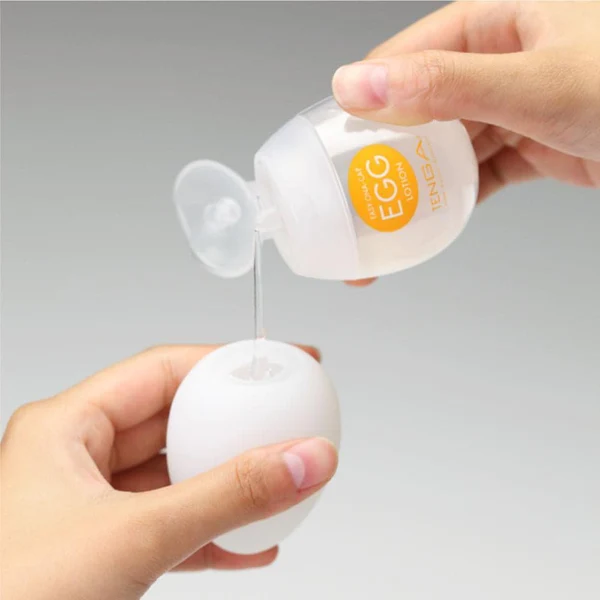 A hand pours Tenga egg lotion from a small bottle into a white egg-shaped container.