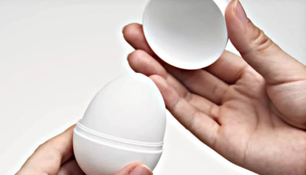 A white egg-shaped masturbator with a ribbed texture is held open by two hands.