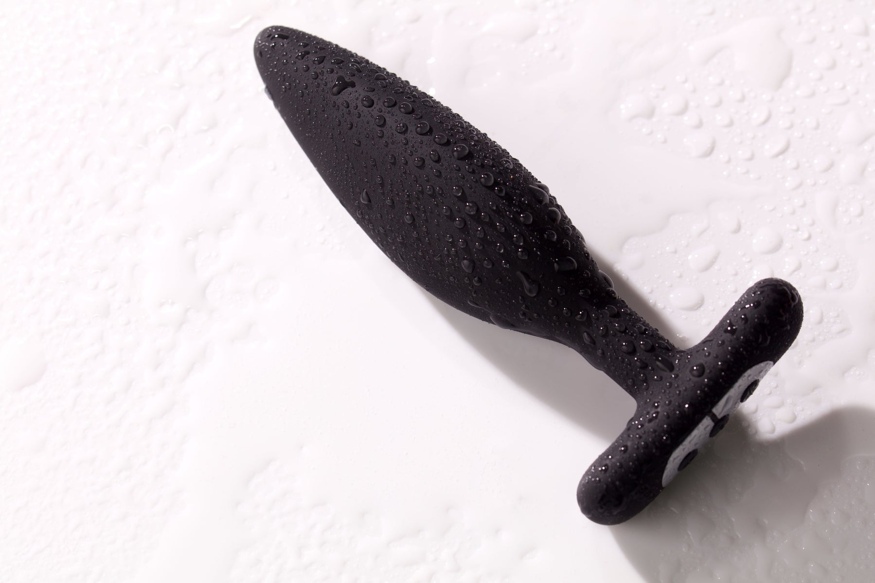 A black vibrating anal plug covered in water droplets on a white surface.