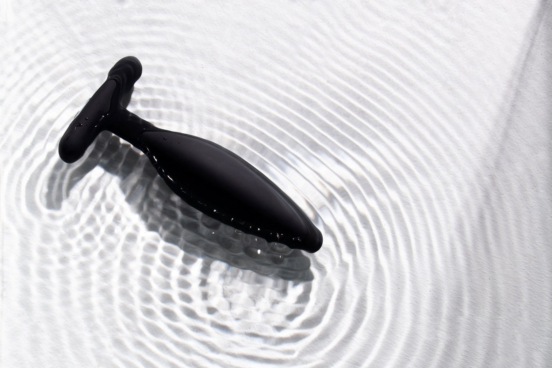 Black silicone vibrating anal plug with a T-shaped handle, resting in rippling water.