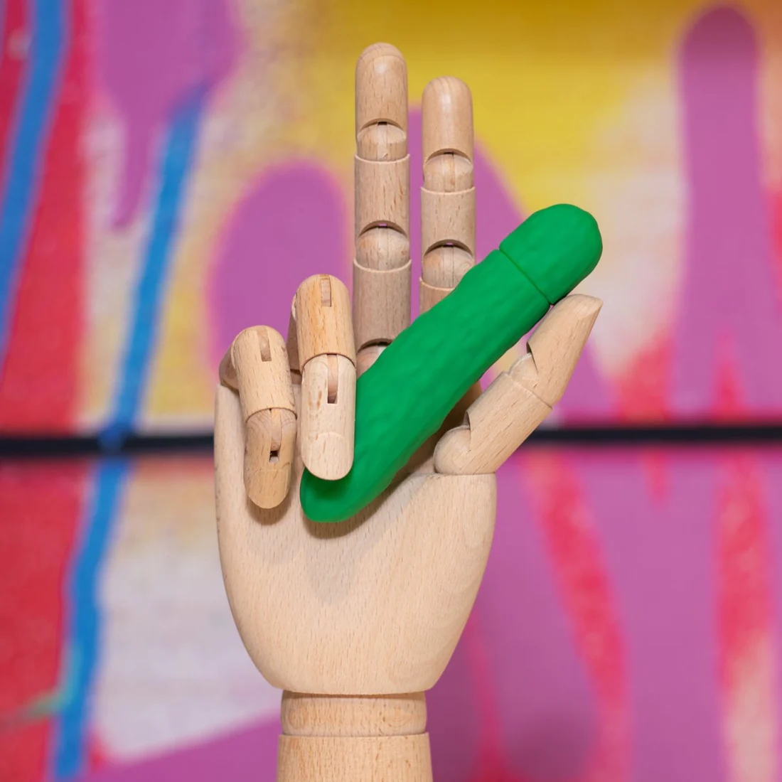 A green pickle-shaped vibrating bullet massager held by a wooden mannequin hand.