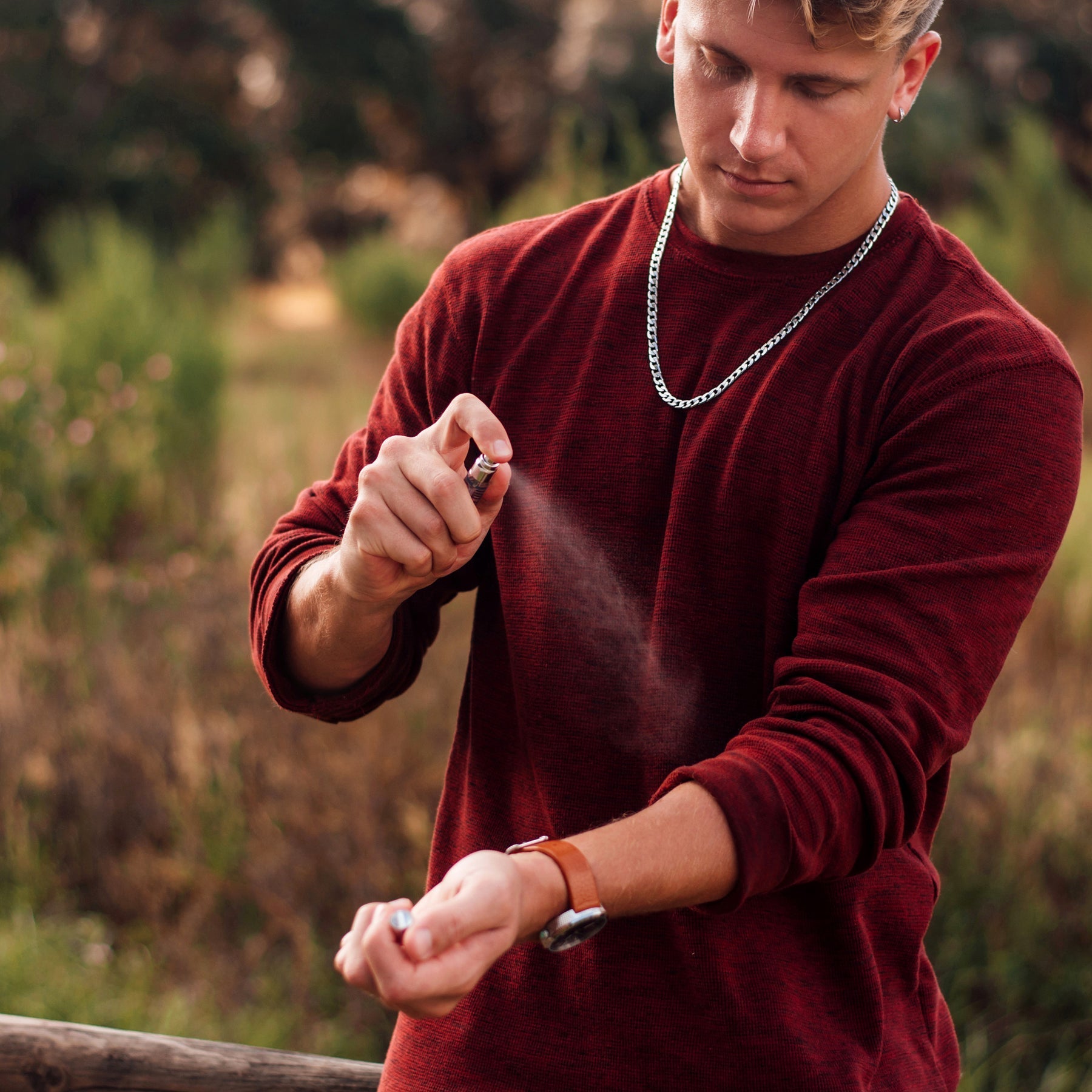 Man spraying cologne on his wrist, wearing a red sweater and silver chain.