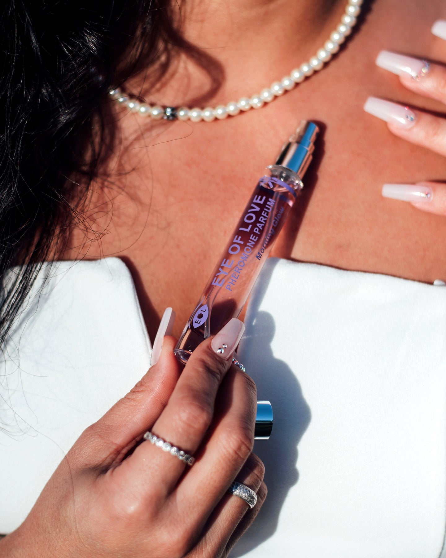 A woman holds a rollerball perfume bottle labeled "Eye of Love" with a pearl necklace.