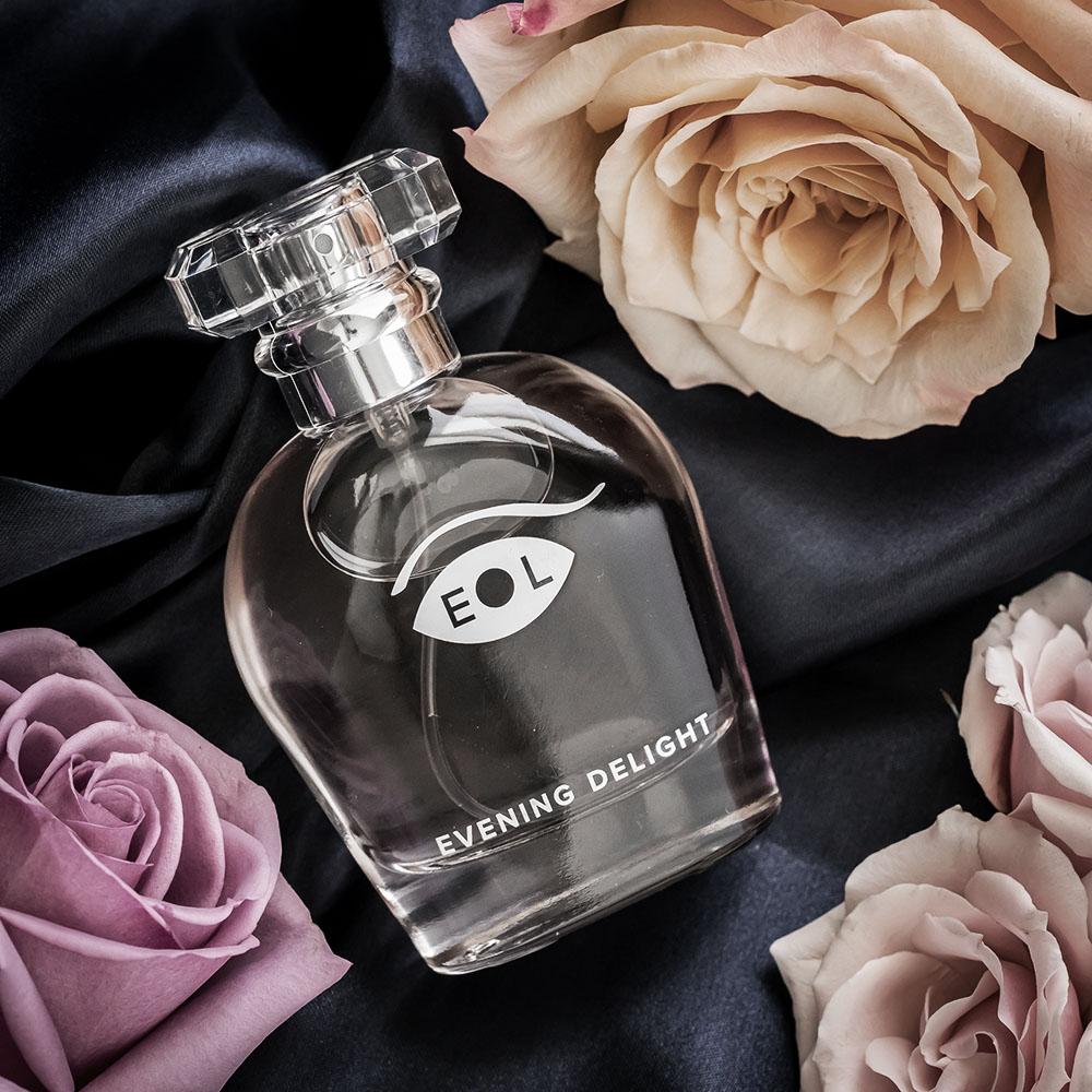 A clear glass bottle of Evening Delight perfume with roses and dark fabric.