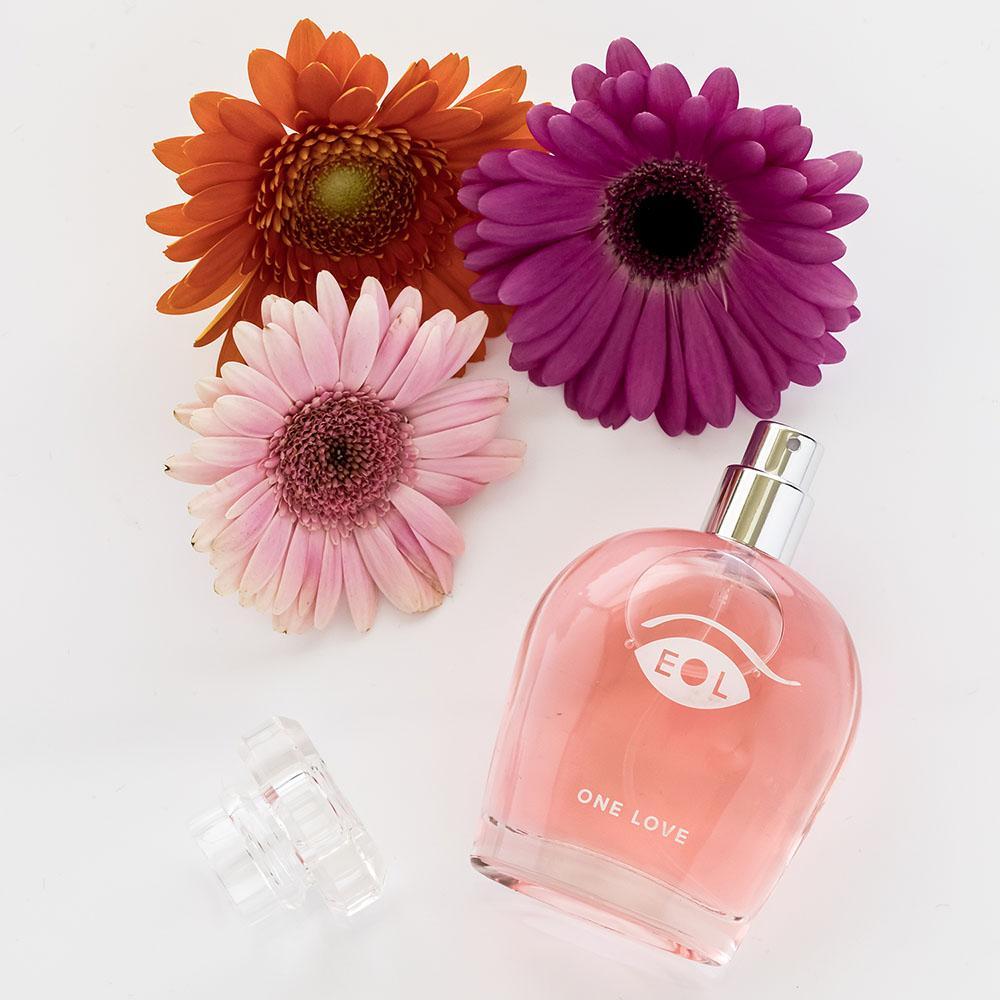 A pink perfume bottle with "ONE LOVE" and an eye logo, next to three gerbera daisies.