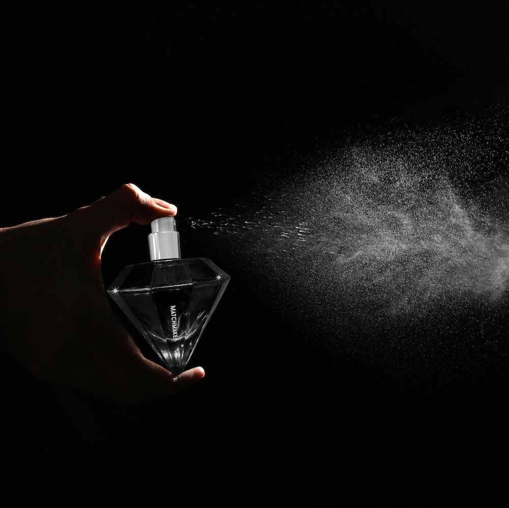 A hand sprays a diamond-shaped bottle of Matchmaker perfume against a black background.