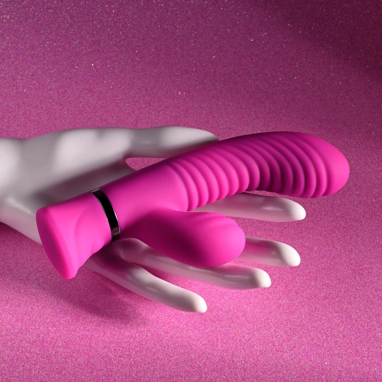 A pink dual silicone vibrator with wavy grooves rests on a white mannequin hand against a sparkly pink background.