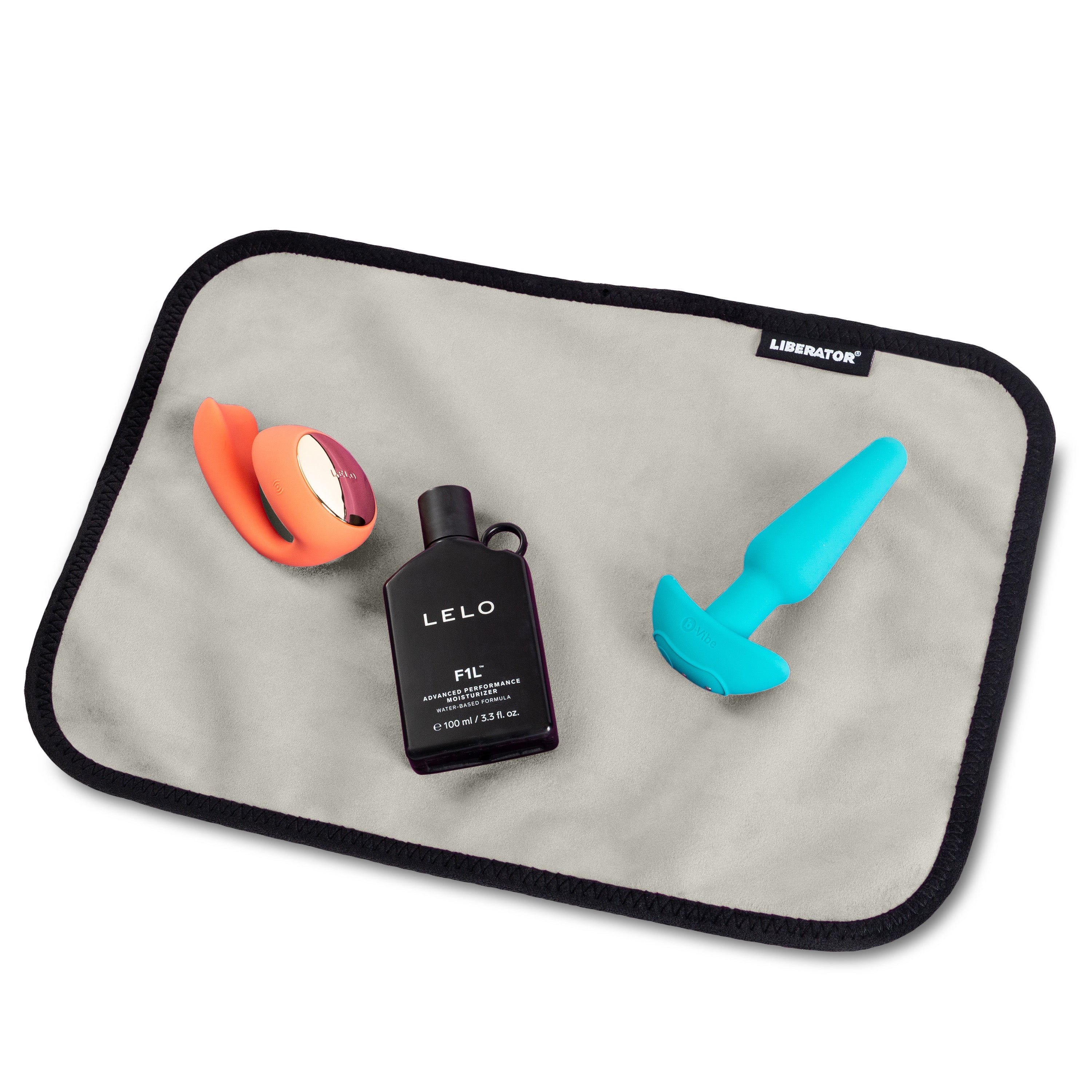 A grey microvelvet mat with black trim, featuring an orange vibrator, a bottle of LELO F1L lubricant, and a teal plug.