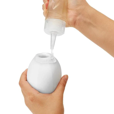 A hand fills a white egg-shaped masturbator with liquid from a clear bottle.