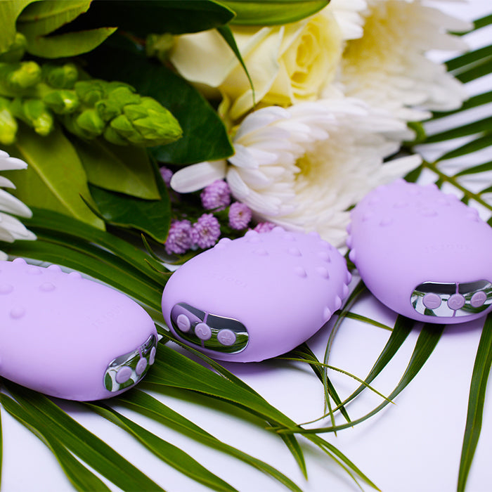 Three purple clitoral vibrators with textured surfaces and silver control buttons rest on green leaves.