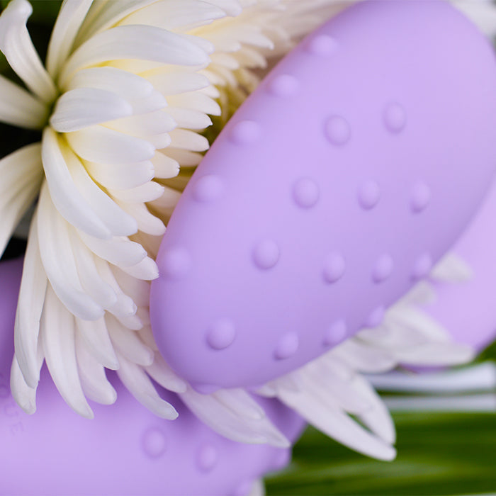 Purple clitoral vibrator with raised dots next to white flower petals.